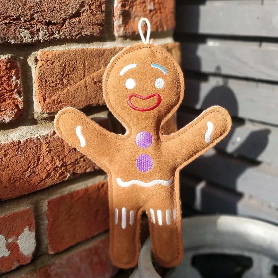 A brown gingerbread person-shaped eco dog toy with suede details stood against a brick wall.