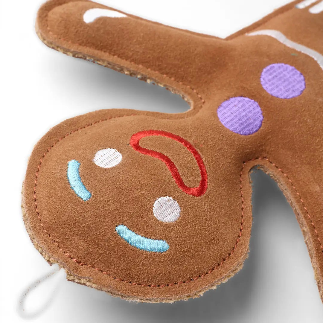 A close up of a brown gingerbread person-shaped eco dog toy with suede details, featuring a red and white face and purple buttons. 